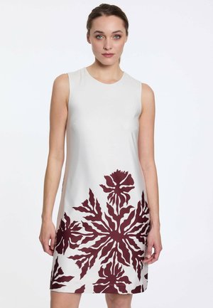 Femme portant une robe blanche sans manches avec un grand imprimé floral rouge foncé le long du bas, debout devant un fond blanc uni.