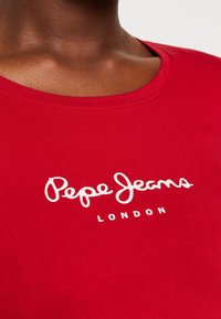 T-shirt in cotone rosso con scollatura rotonda, caratterizzato da testo bianco che recita "Pepe Jeans London." Texture liscia, senza motivi visibili.