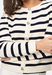 Cardigan tricoté crème avec des rayures horizontales bleu marine, présentant des boutons ronds métalliques, des poignets côtelés et une texture lisse.