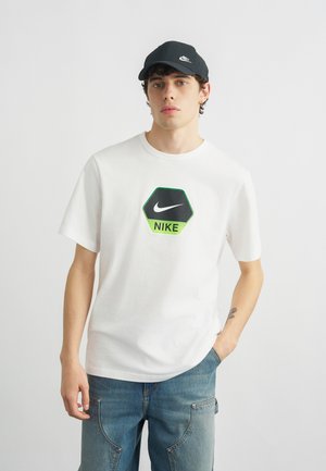 Mladić u bijeloj Nike majici s heksagonalnim logotipom, plavim trapericama i crnoj Nike kapa, stoji s jednom rukom u džepu.