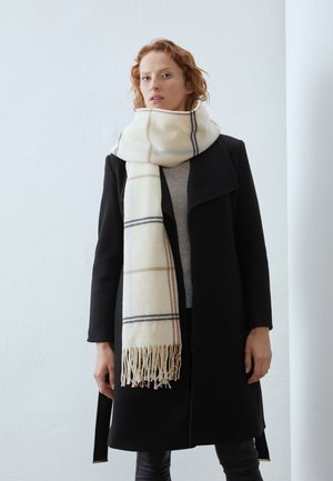 Femme aux cheveux roux porte un manteau noir et une grande écharpe blanche à carreaux avec des franges enroulée autour de son cou sur un fond blanc uni.