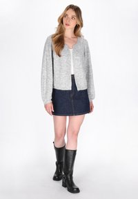 Femeie în picioare, îmbrăcată cu un cardigan gri, un tricou alb, o fustă din denim închis și cizmă neagră până la genunchi, privind ușor spre stânga.