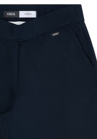 Pantalon bleu marine avec plis à l'avant, poches latérales et une petite étiquette métallique de la marque "Cecil" près de la ceinture à l'intérieur.