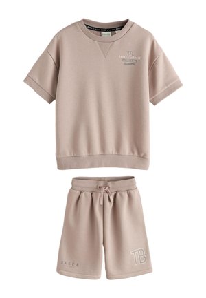 Dreng med krøllet hår iført beige oversized T-shirt og shorts, hvide sneakers, står vendt mod en ensfarvet lys grå væg.