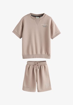 Dreng med krøllet hår iført beige oversized T-shirt og shorts, hvide sneakers, står vendt mod en ensfarvet lys grå væg.