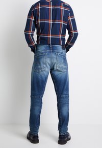 Hombre vistiendo jeans azules desgastados, camisa de cuadros en navy y naranja metida por dentro, y zapatos oscuros, de pie frente a una pared blanca lisa.