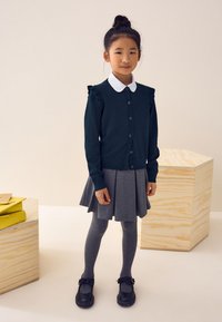 Granatowy kardigan z falbankami, biały kołnierzyk, szara plisowana spódnica, szare rajstopy i czarne buty z kokardkami. Prosty, teksturowany wzór.