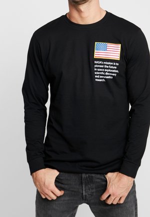 Mężczyzna w czarnej koszulce z długim rękawem z flagą USA i napisem dotyczących misji NASA na piersi, ręce blisko kieszeni, na jednolitym jasnym tle.