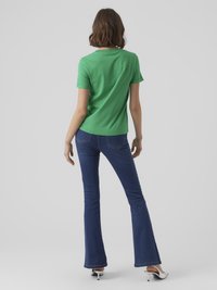 Moteris su pečių ilgio rudais plaukais, vilkinti žalią trumpų rankovių marškinėlį, mėlynus plačius džinsus ir baltus aukštakulnius, stovinti prieš pilką fono.