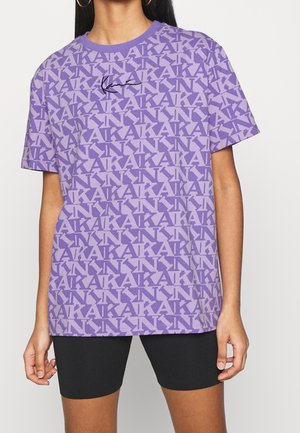 Kobieta ubrana w fioletową koszulkę z wzorem, na której powtarza się tekst "KARL" oraz czarny nadruk z podpisem, w zestawieniu z czarnymi szortami rowerowymi.