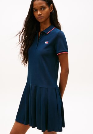 Frau trägt ein marineblaues Polokleid mit rosa Besatz am Kragen und an den Ärmeln, steht vor einem schlichten weißen Hintergrund.