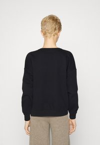 Persona con cabello corto rubio, vistiendo una camiseta de manga larga negra y pantalones beige, de pie de espaldas frente a una pared blanca.