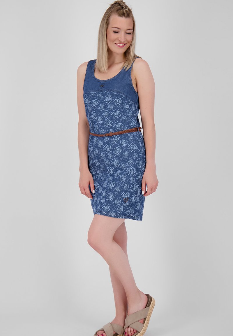 Vrouw in een mouwloos blauw patroonjurk met een bruin gevlochten riem en beige wedge sandalen, staand tegen een effen achtergrond.