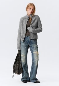 Γκρι πλεκτό ζακέτα με ραβδωτό στρίφωμα, φορεμένη πάνω από μια ριγέ μπλούζα. Ξεφτισμένα, φαρδιά τζιν σε μπλε απόχρωση με ανοιχτούς λεκέδες βαφής και μια μαύρη τσάντα.