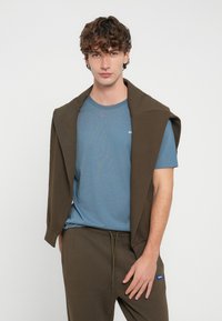 Hellblaues T-Shirt mit kleinem Logo, darüber ein dunkelbrauner Pullover, der über die Schultern gelegt ist, kombiniert mit passenden braunen Jogginghosen.