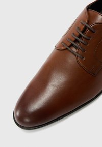 Chaussure habillée en cuir marron avec un bout arrondi élégant, des coutures décoratives et cinq œillets pour les lacets, présentant une finition lisse et brillante.
