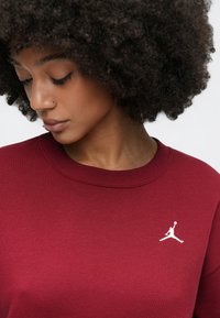 Κόκκινο φούτερ με ραβδωτό λαιμόκοψη, με λευκό λογότυπο Jumpman στο αριστερό στήθος. Το ύ φασμα φαίνεται μαλακό με υφή.