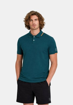 Uomo con capelli ricci che indossa una polo verde acqua con dettagli arancioni e pantaloncini neri, in piedi con le mani nelle tasche davanti a uno sfondo bianco.