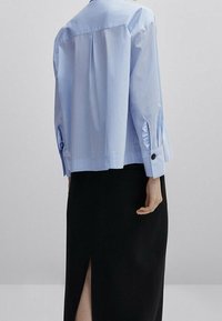 Chemise bleu clair à boutons avec une coupe décontractée, des plis au dos et des manches retroussées. Associée à une jupe noire dotée d'une fente latérale.