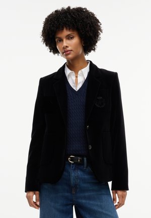 Femme aux cheveux bouclés portant un blazer noir, un gilet bleu marine, une chemise blanche à col, un jean bleu et une ceinture noire.