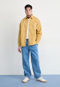 Dzeltena denima krekls ar priekšējo kabatu, balts T-krekls zem tā, zilas sporta bikses un baltas tenisa kurpes ar gumijas zoli. Vienkārša fona.