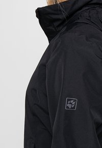 Svart vattentät jacka med hög krage, med ett tassavtryck-logotyp på ärmen och strukturerad tyg för väderbeständighet.