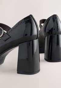 Black patent leather Mary Jane heels with chunky block heels and perforated detailing near the strap and edges on a light background.
