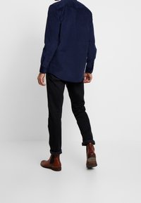 Laivastonsininen velourpaita pitkillä hihoilla, napilliset kalvosimet ja klassinen kaulus, yhdistetty mustiin housuihin ja ruskeisiin nilkkureihin.