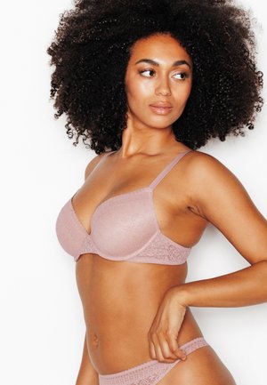 Donna con capelli ricci indossa un completo coordinato di reggiseno e biancheria intima in pizzo rosa chiaro, guarda a sinistra con la mano appoggiata sul fianco.