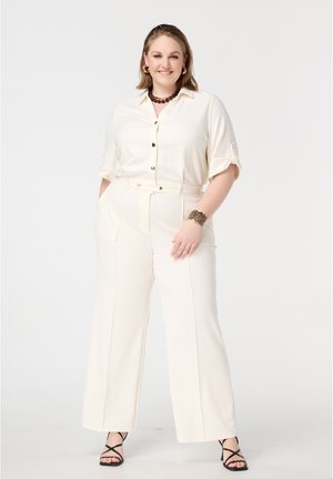 Femme souriante, portant une chemise crème à boutons et un pantalon taille haute crème, avec des talons noirs à lanières, accessoirisée d'un collier et d'un bracelet.