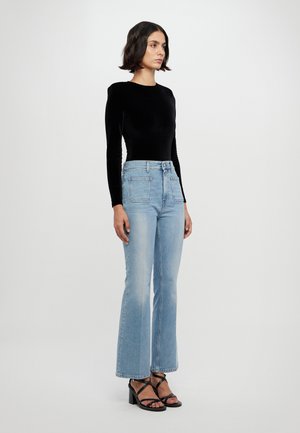 Svart fløyelsbluse med lange ermer, kombinert med lyseblå, høytliv brunfargede jeans med utvidede ben. Modellen har på seg svarte, stilige hæler. Enkelt, rent design.
