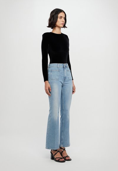 Haut à manches longues en velours noir associé à un jean flare taille haute bleu clair, et des sandales à talons noires avec lanières. Design simple, détails minimalistes.