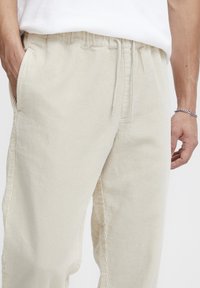 Pantalon en velours côtelé beige à rayures verticales, doté d'une taille élastique et de poches latérales, photographié de la taille aux pieds.