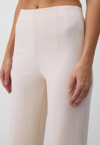 Helle rosa Hose aus glattem Stoff, mit hoher Taille, zwei Vorderfalten und weitem Bein. Texturierte Oberfläche verleiht subtile Details.