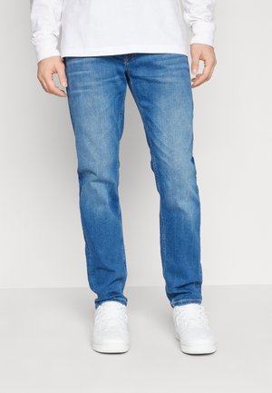 Homme portant un jean slim bleu clair, une chemise blanche à manches longues et des baskets blanches, debout devant un fond uni.