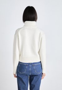 Hvit strikket genser med høy ribbestrikket krage, cropped passform og ribbestrikket hem. Kombinert med blå denimjeans med baklommer.