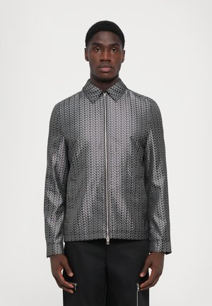 Metallic jacket featuring a geometric pattern in silver and black, with a zip front, pointed collar, and long sleeves.