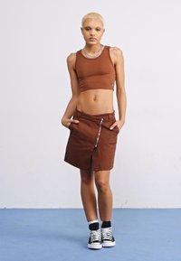 Top corto marrón y falda hasta la rodilla con cierre de cremallera, ambos de algodón. La modelo lleva zapatillas negras y blancas; fondo liso.