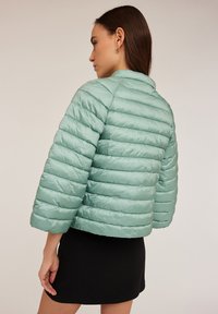 Jasnozielona kurtka puchowa o poziomym pikowanym wzorze, z kołnierzem i rękawami do trzech czwartych. Noszona z czarną spódnicą.