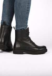 Černé kožené šněrovací boty na noze s modrými džíny, zobrazené z bočního a zadního pohledu na světlém pozadí.