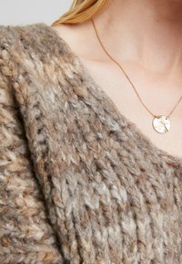 Närbild av en person som bär en tjockstickad brun tröja och ett tunt guldhalsband med en rund, texturerad berlock.