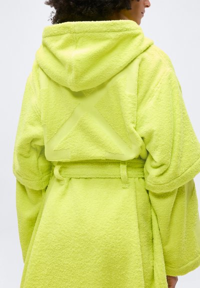Personne portant une robe à capuche texturée d'un vert citron vif avec de larges manches et une ceinture nouée à la taille, de dos au spectateur.