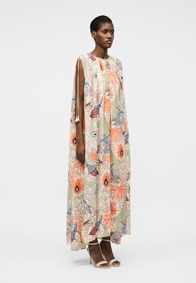 Γυναίκα που στέκεται σε ένα χαλαρό, μακρύ φόρεμα χωρίς μανίκια με πολύχρωμο λουλουδένιο μοτίβο και λεπτομέρειες δεσίματος στους ώμους, φορώντας λευκά σανδάλια με ψηλό τακούνι.