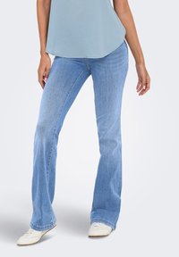 Η γυναίκα φοράει ανοιχτό μπλε τζιν bootcut, λευκά αθλητικά παπούτσια και ένα ανοιχτό μπλε αμάνικο τοπ, στέκεται μπροστά σε ένα απλό φωτεινό φόντο.