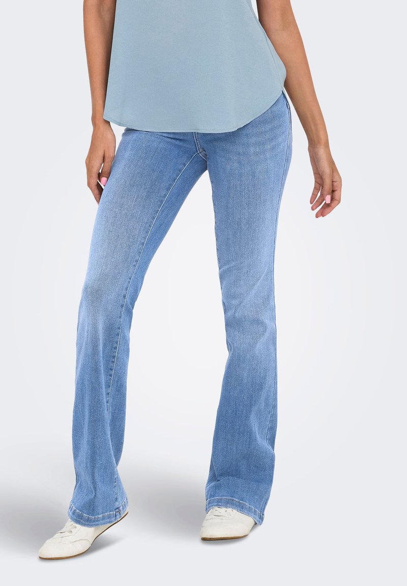 Η γυναίκα φοράει ανοιχτό μπλε τζιν bootcut, λευκά αθλητικά παπούτσια και ένα ανοιχτό μπλε αμάνικο τοπ, στέκεται μπροστά σε ένα απλό φωτεινό φόντο.