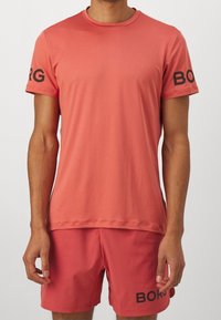 T-shirt de sport corail à manches courtes avec encolure ronde ; présente le logo "BORG" en noir sur les manches. Tissu doux et léger, coupe ajustée.