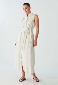 Knee-length, bezrukávový, zapínaný na gombíky ľanový šaty v off-white farbe s opaskom v páse a vrstveným lemom. Obsahuje golier a jemnú textúru.