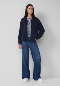 Donna che indossa una giacca blu navy con zip, una blusa a fantasia, jeans larghi blu e sneakers bianche contro uno sfondo grigio uniforme.