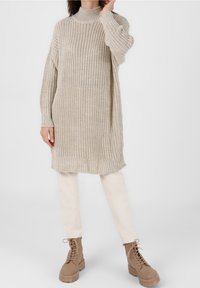 Robe pull beige en tricot avec un col haut, des poignets côtelés et un ourlet à franges. Associée à un pantalon clair et des bottes marron à lacets.