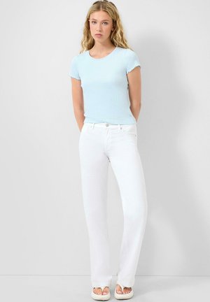 Młoda kobieta z długimi blond włosami, ubrana w jasnoniebieską bluzkę z krótkim rękawem, białe spodnie z wysokim stanem i białe sandały z otwartymi palcami, stoi na tle białej ściany.